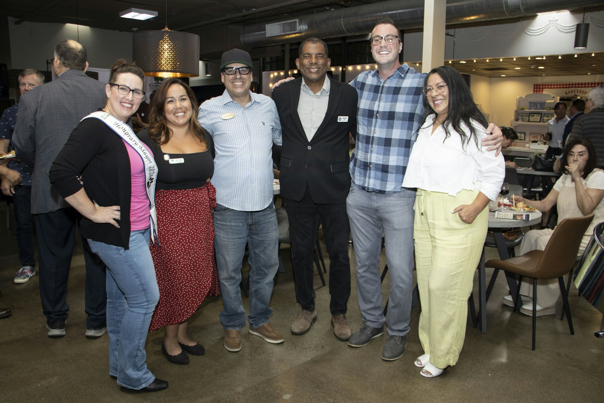 A group of six people posing together, smiling in an indoor setting with others in the background.