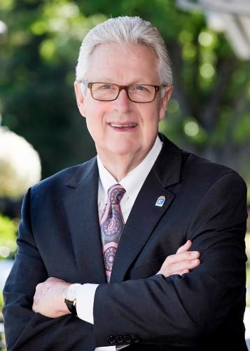 A man in a suit and tie, wearing glasses, smiling with arms crossed outdoors.