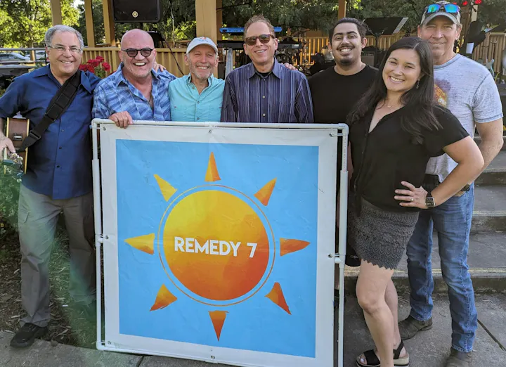 A group of seven people poses with a sign for "REMEDY 7," likely a music or performance group, outdoors in a casual setting.