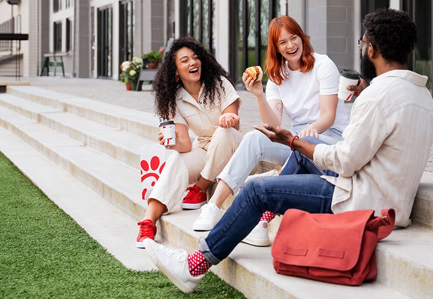 Three people sitting on steps, laughing and enjoying drinks and snacks outdoors.