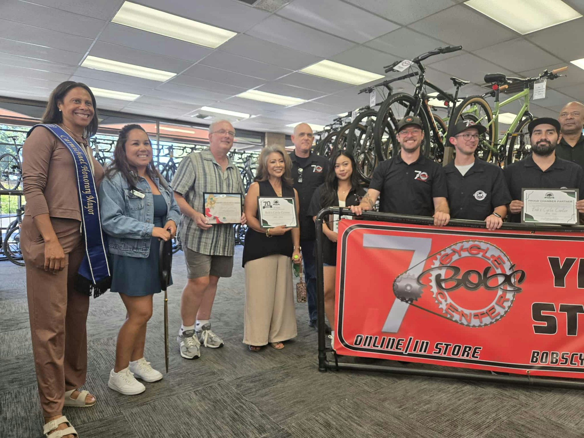 A group of people posing in a bicycle shop, holding certificates, with a "Bob's Cycle Center" banner in front of them.