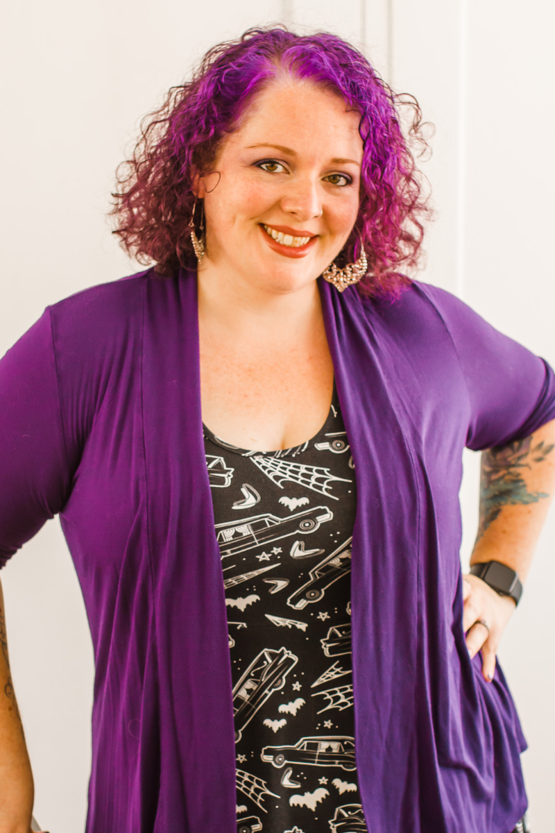 A woman with purple hair and cardigan smiling at the camera.