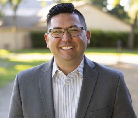 A smiling man in a suit with glasses, outdoors.