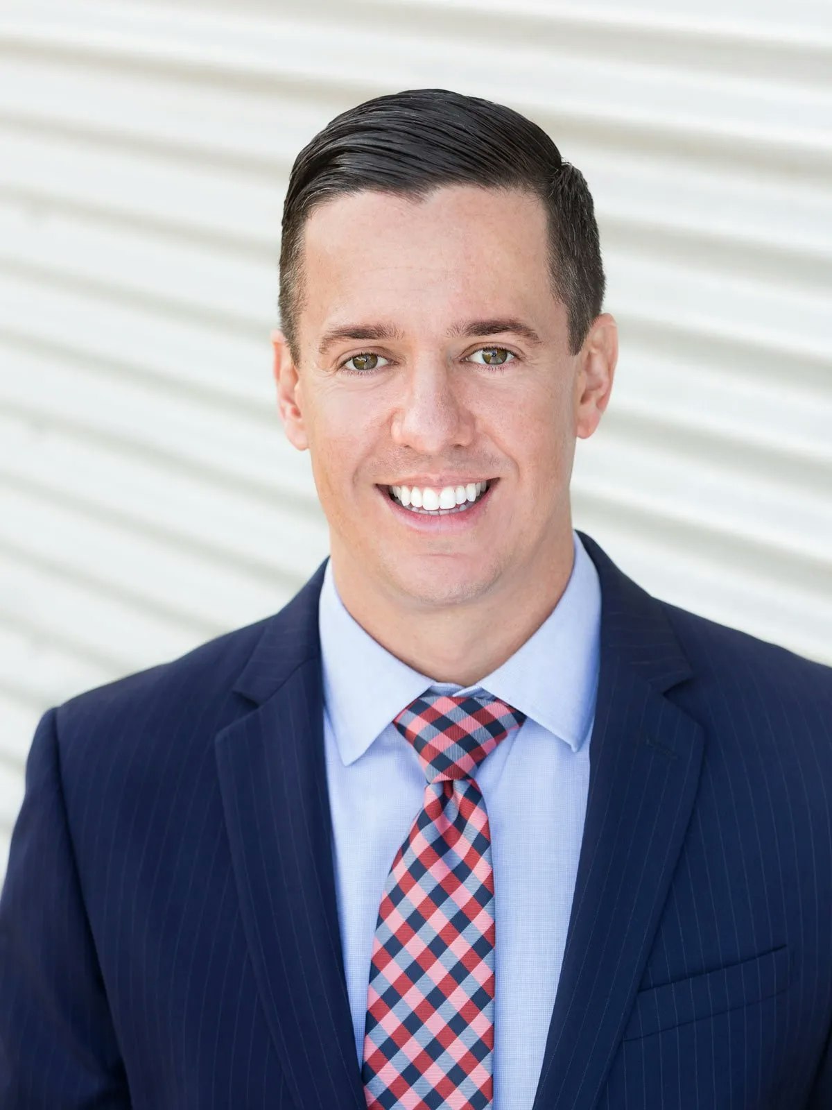 A person in a suit and checkered tie smiles against a striped background.