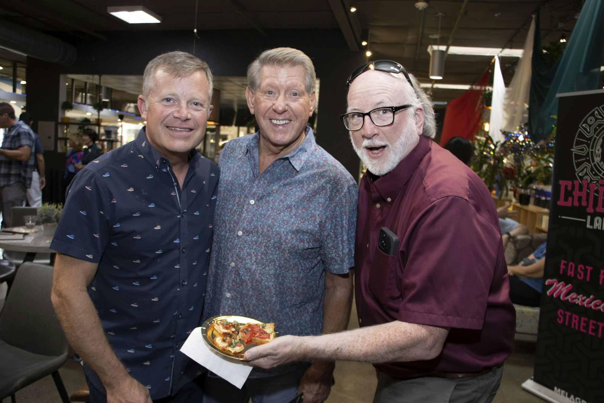 Three men smiling indoors, one holding a plate of food.