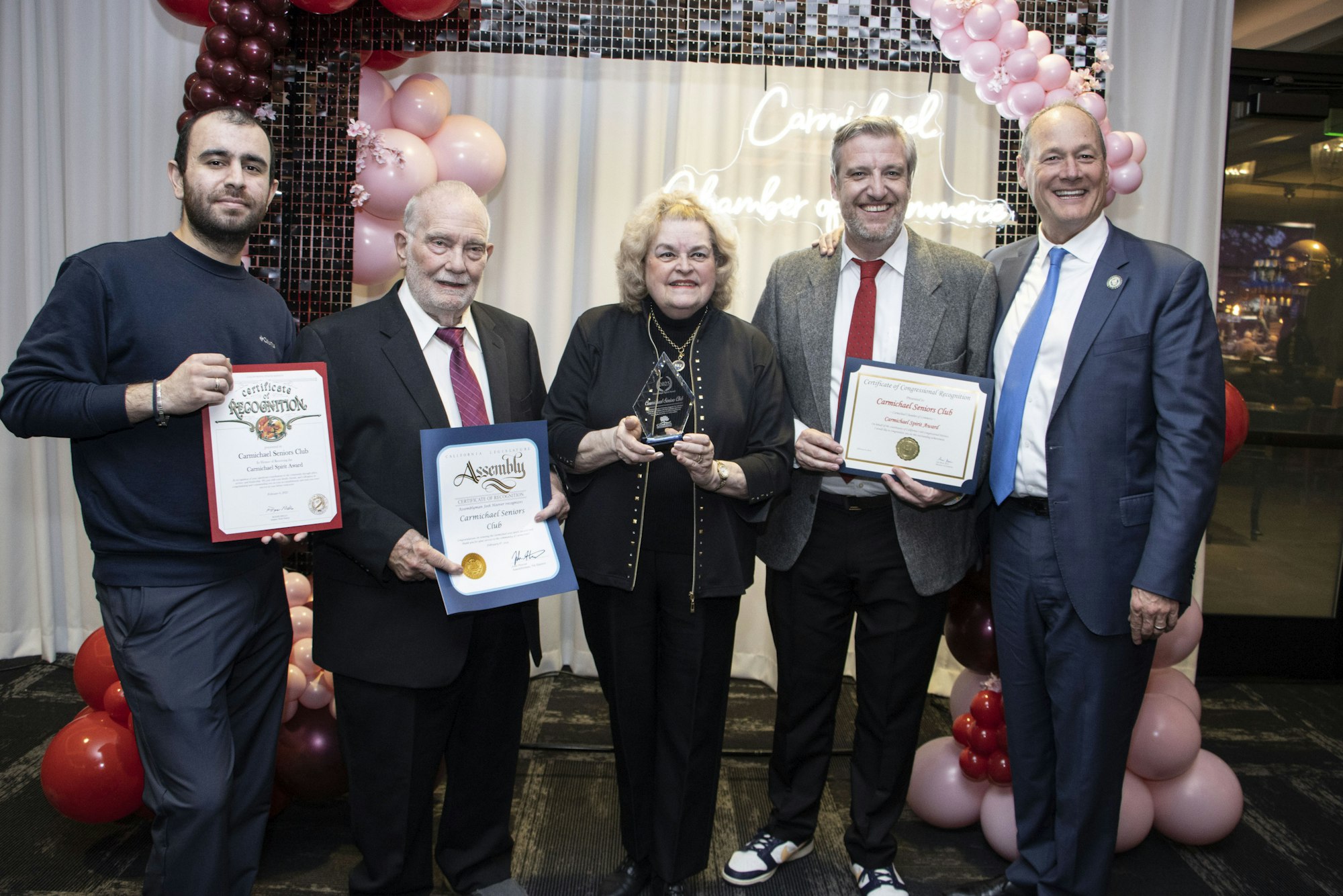 Group of five people holding certificates and an award, with a balloon arch in the background.