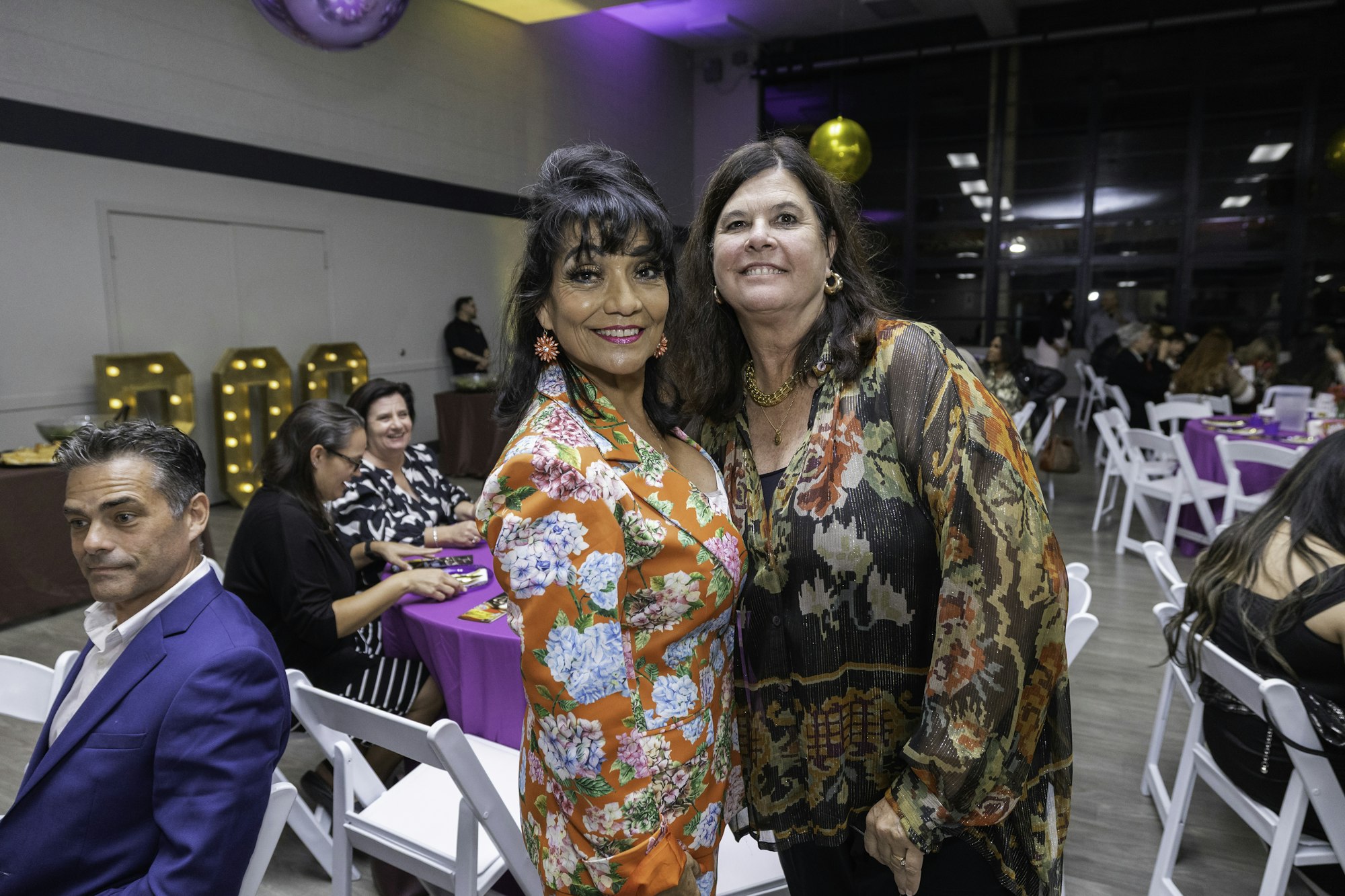 Two women smiling at a festive event, surrounded by other people seated at tables.
