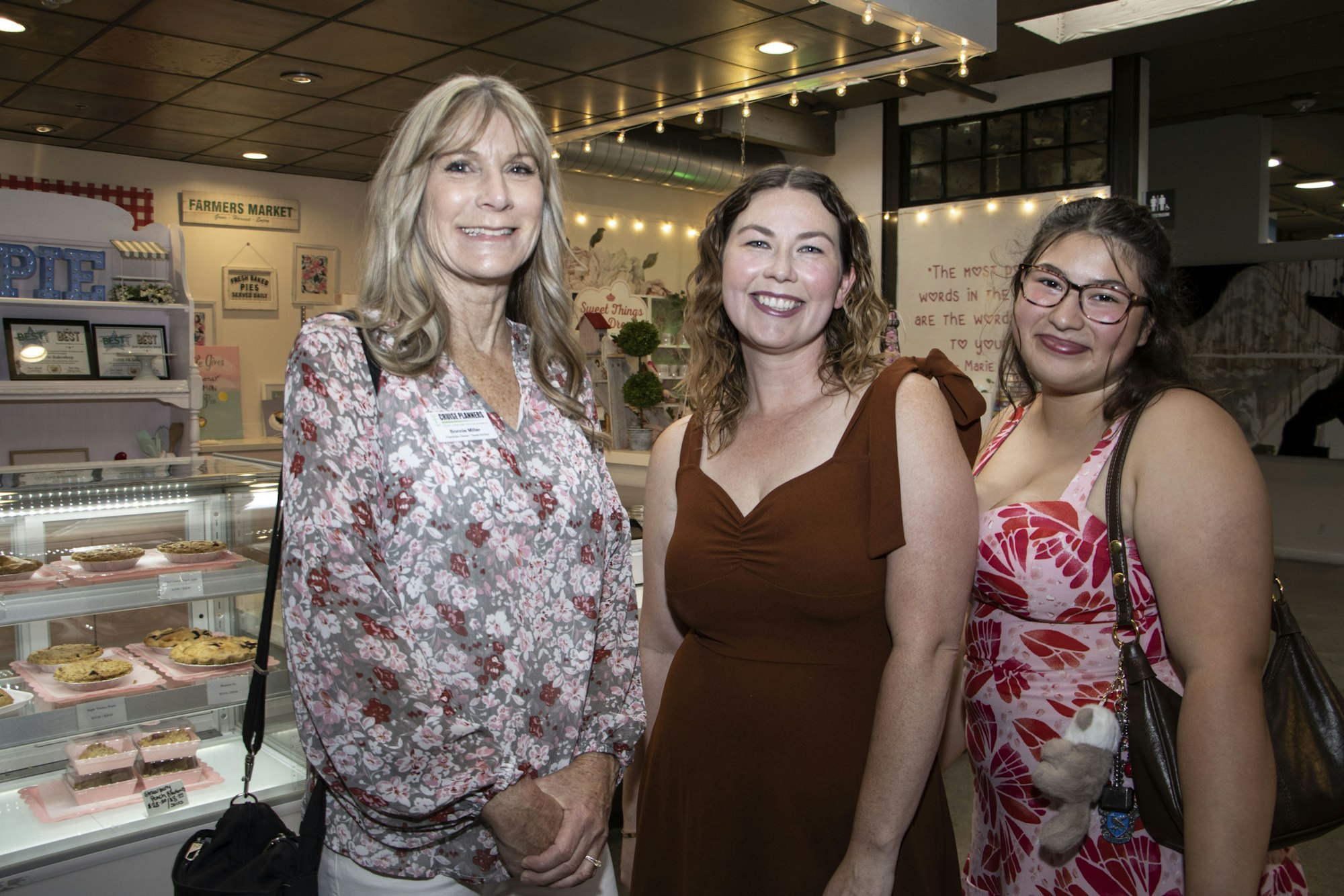 Three women smiling inside a bakery, with pies in a display case.