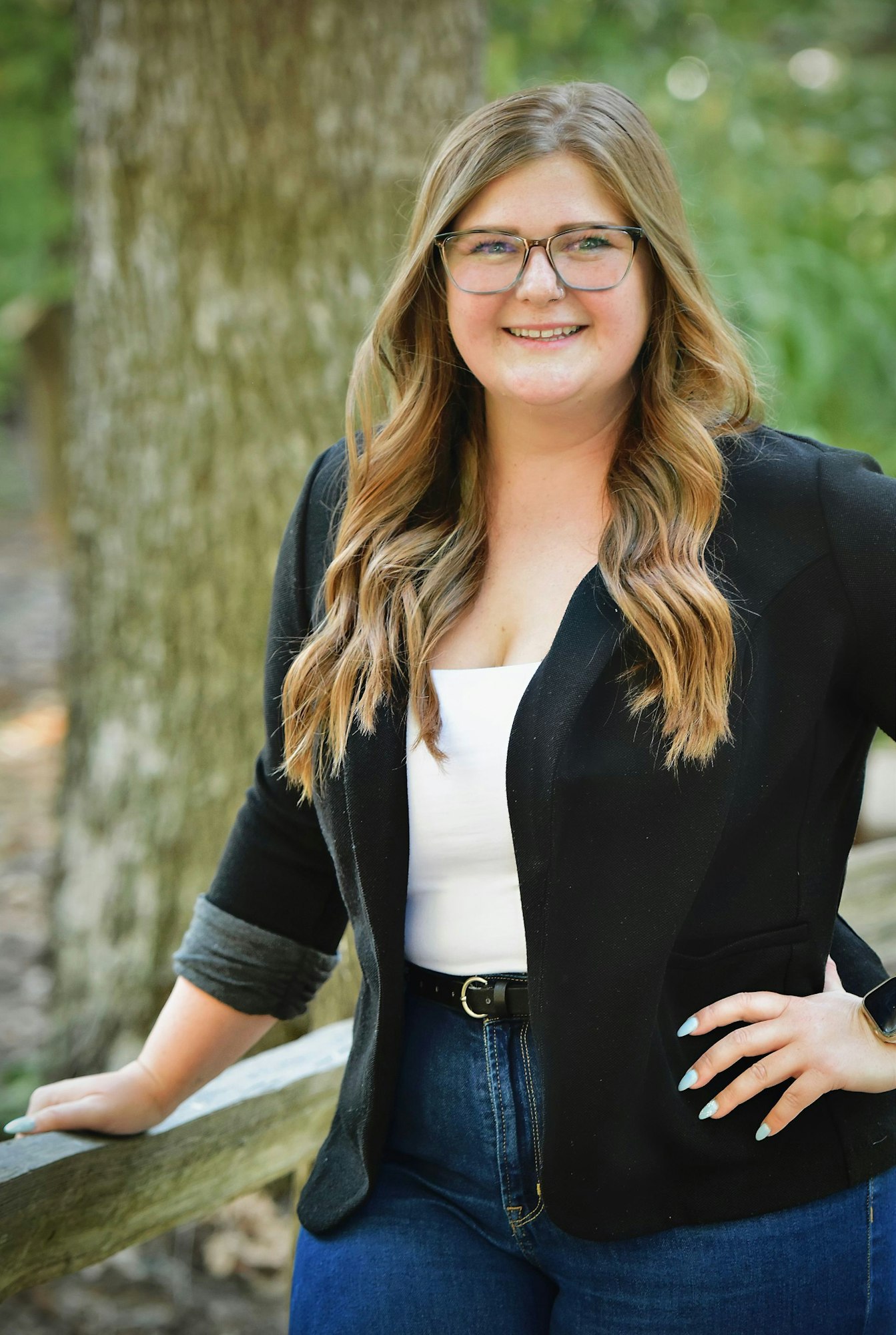 A smiling woman with glasses, wearing a blazer and jeans, leaning on a fence outdoors.