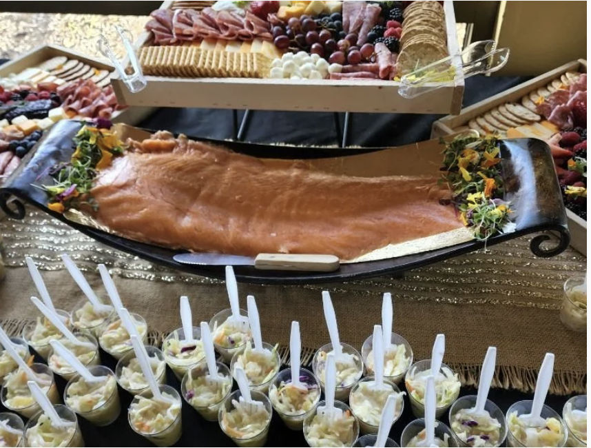 The image features a lavish spread of smoked salmon, assorted meats, cheeses, fruits, and dessert cups on a decorative table.