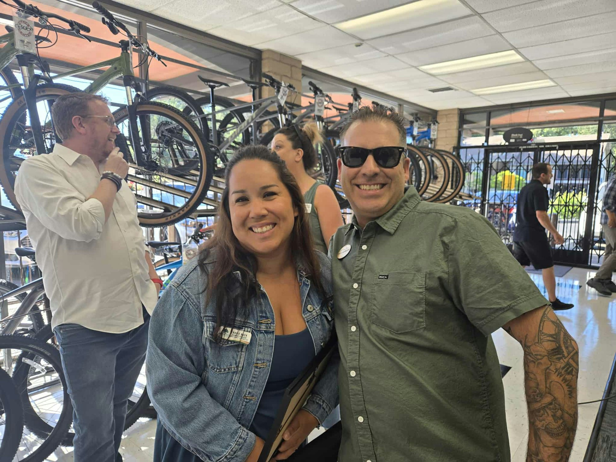 A man and woman smiling in a bike shop, with people and bicycles in the background.