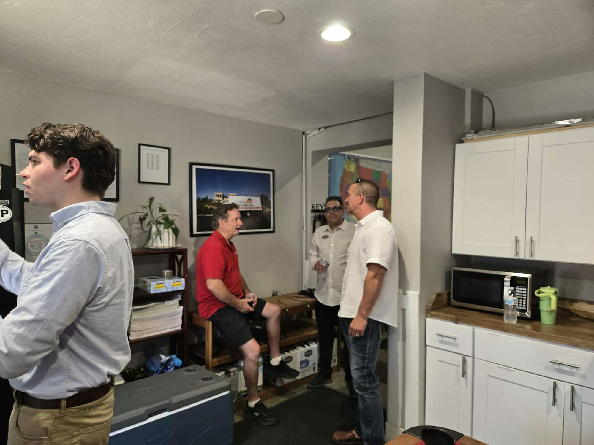 A group of four men socializing in a room with a microwave, cabinets, and framed pictures.