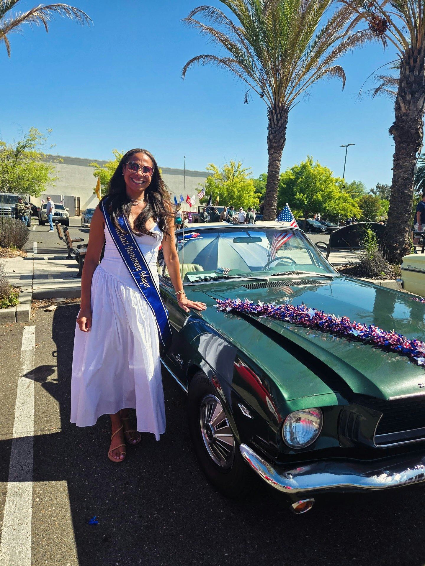 A woman in a white dress with a sash stands by a green classic car adorned with flag-themed decorations, under palm trees.