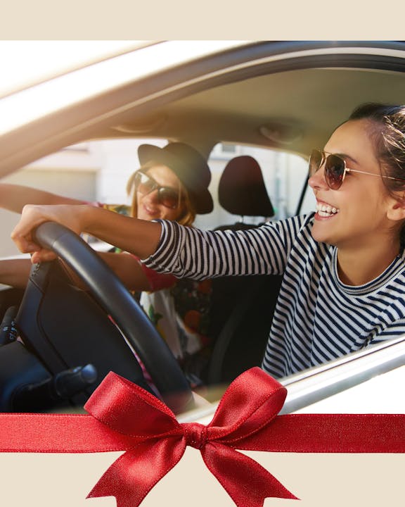 Two friends are enjoying a ride in a car, smiling and having fun, with a festive red bow graphic at the bottom.