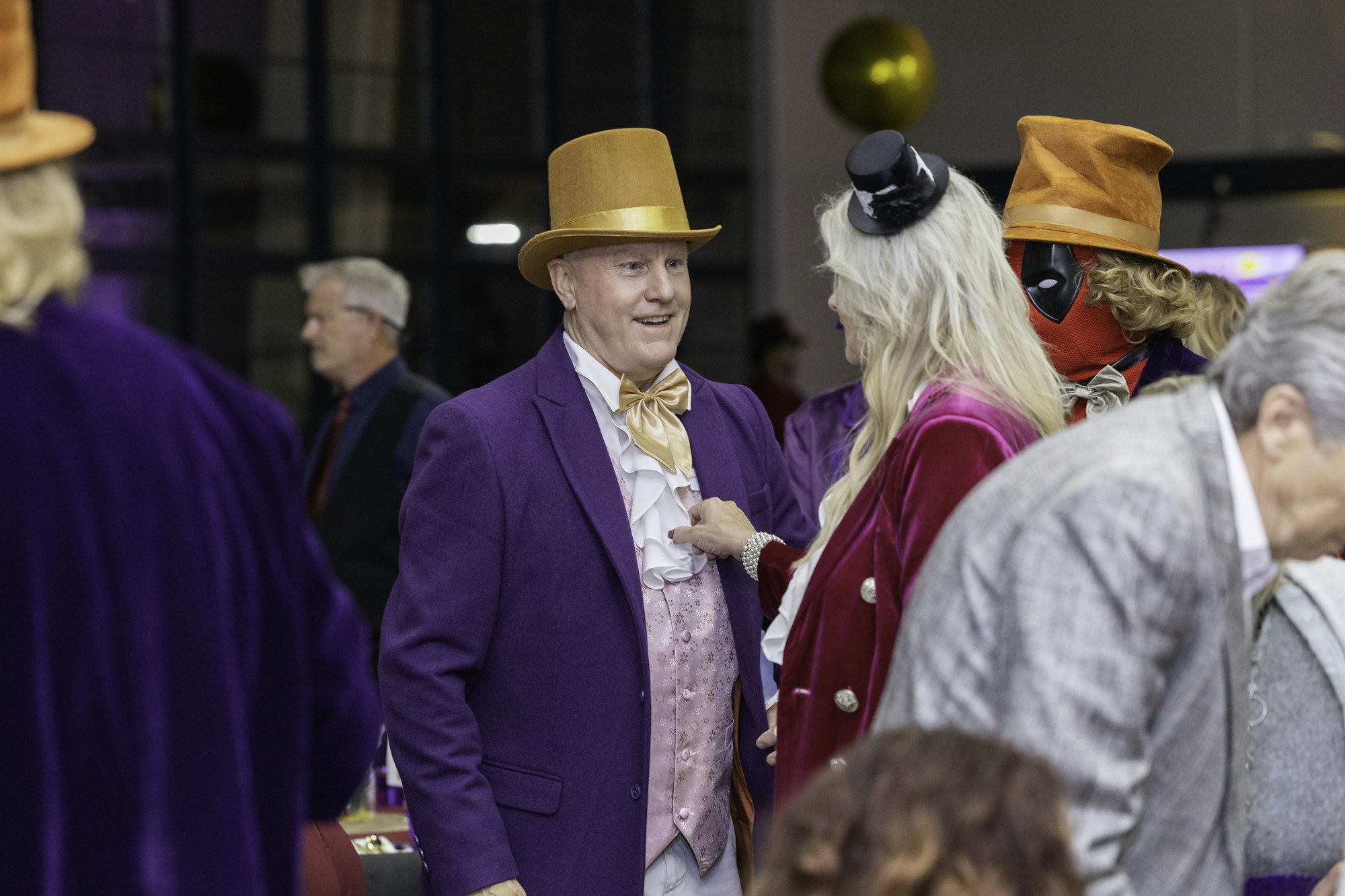 People dressed in elegant, colorful costumes, some with top hats, at an event or gathering indoors.