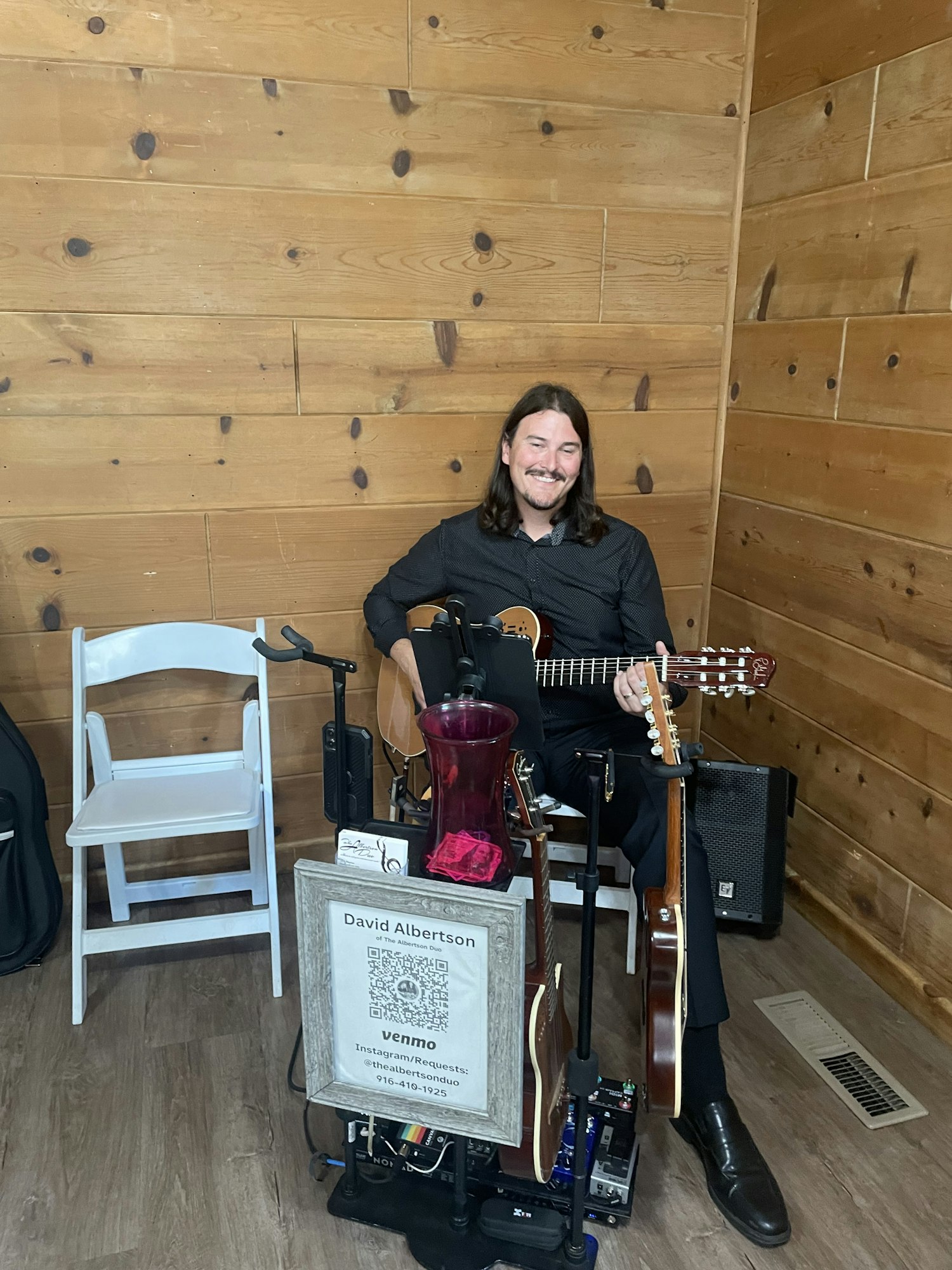 A musician smiles while playing guitar, sitting beside equipment and a sign with payment info, on a wooden background.