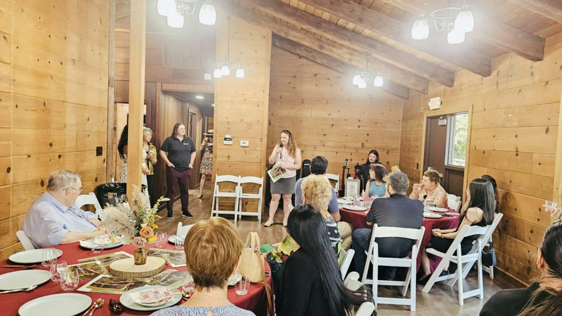 A group of people gathered in a wooden room for a meal, with one person standing and speaking to the group.