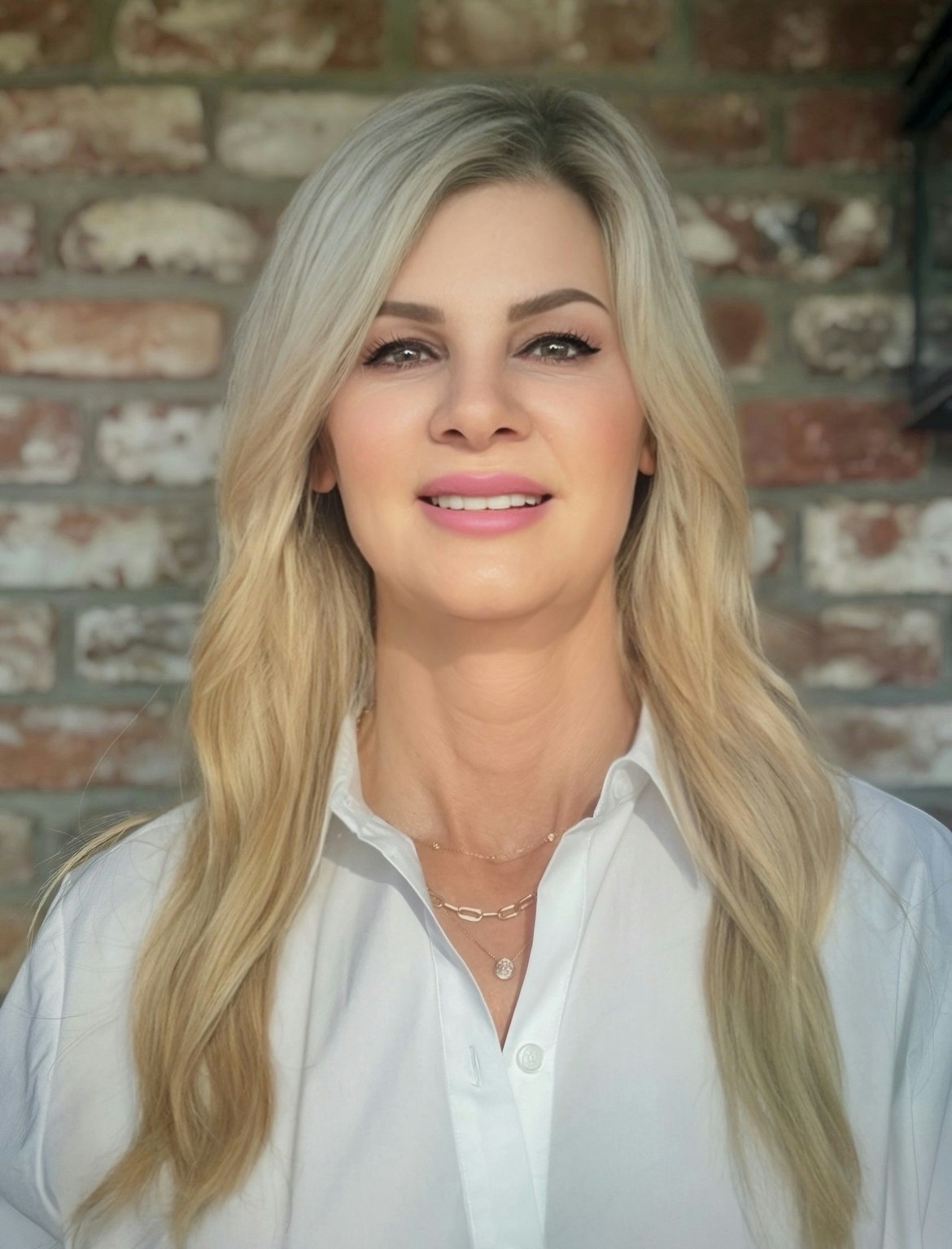 A person with blonde hair wearing a white shirt, smiling in front of a brick wall.