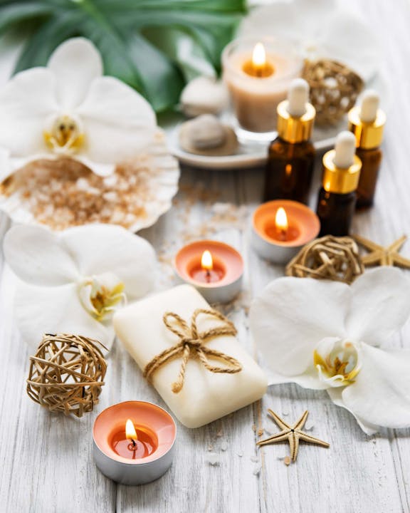 Spa scene with candles, essential oils, soap wrapped in twine, seashells, and white orchids on a wooden surface.