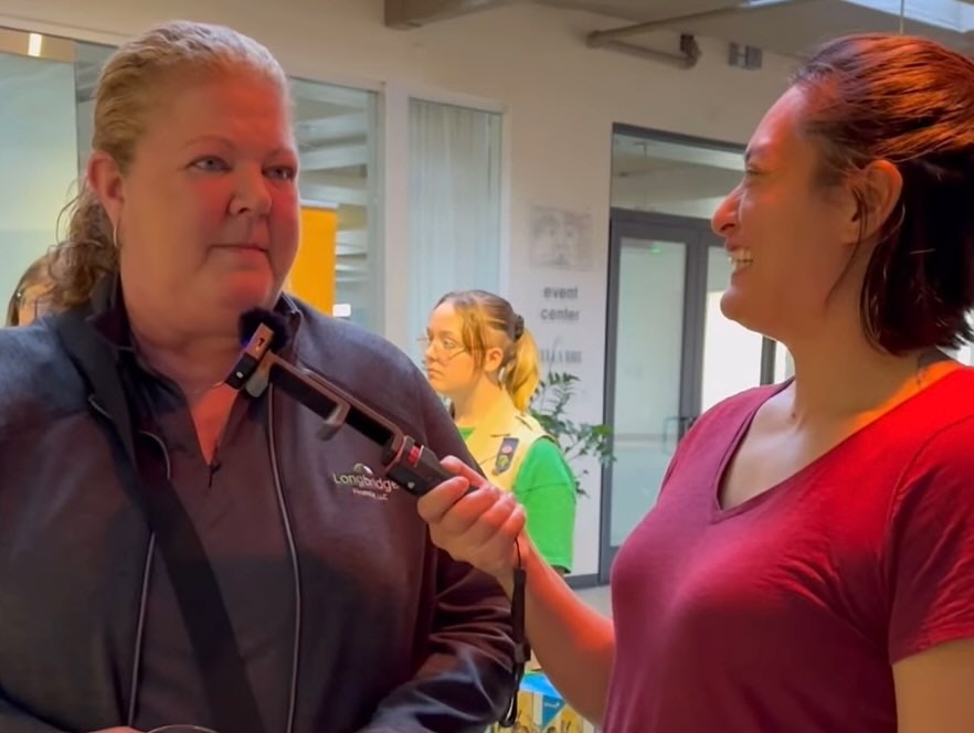 Two people indoors: one being interviewed while holding a selfie stick with a microphone; another person is in the background.