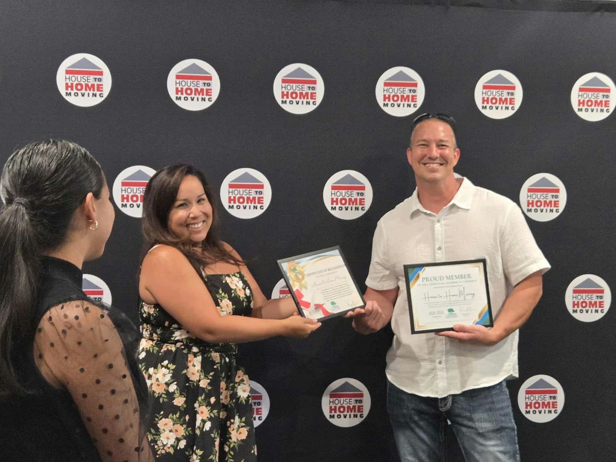 Three people at a "House to Home Moving" event, smiling, with two of them holding certificates.