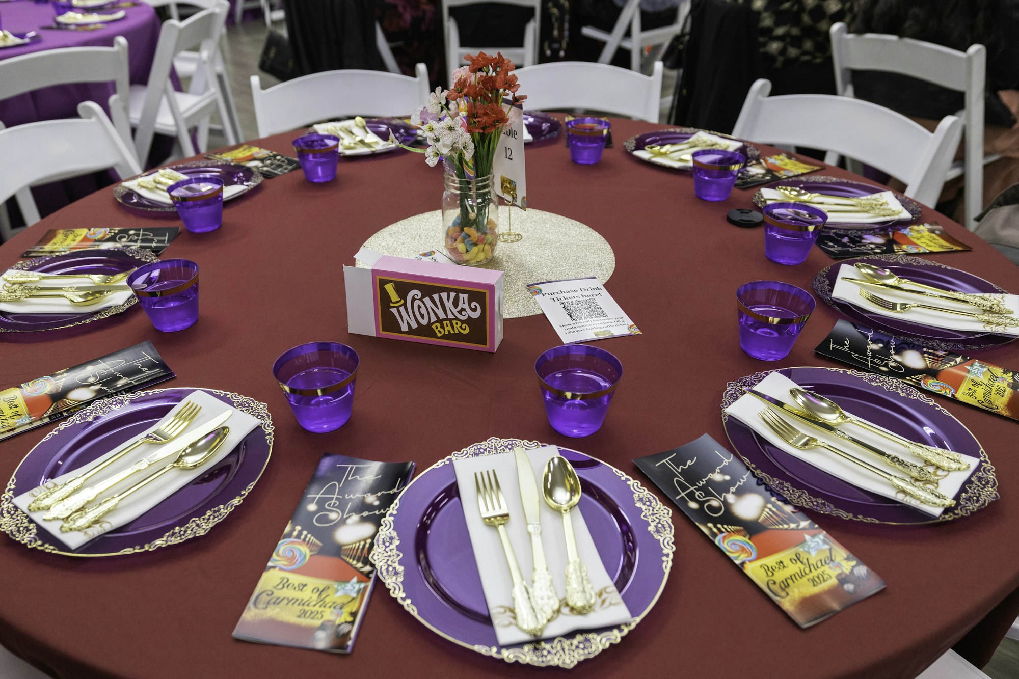 A round table with purple plates, gold cutlery, Wonka Bar decor, purple cups, brochures, and a flower centerpiece.