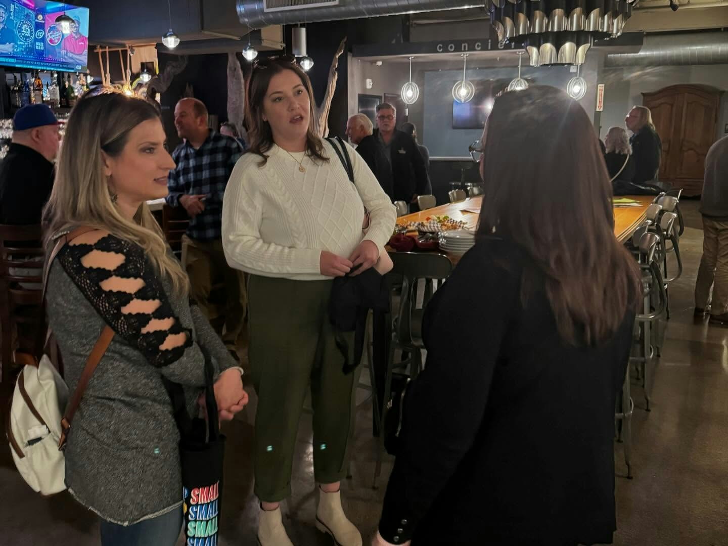 A group of people talking in a bar or restaurant setting with food, drinks, and a TV screen visible.