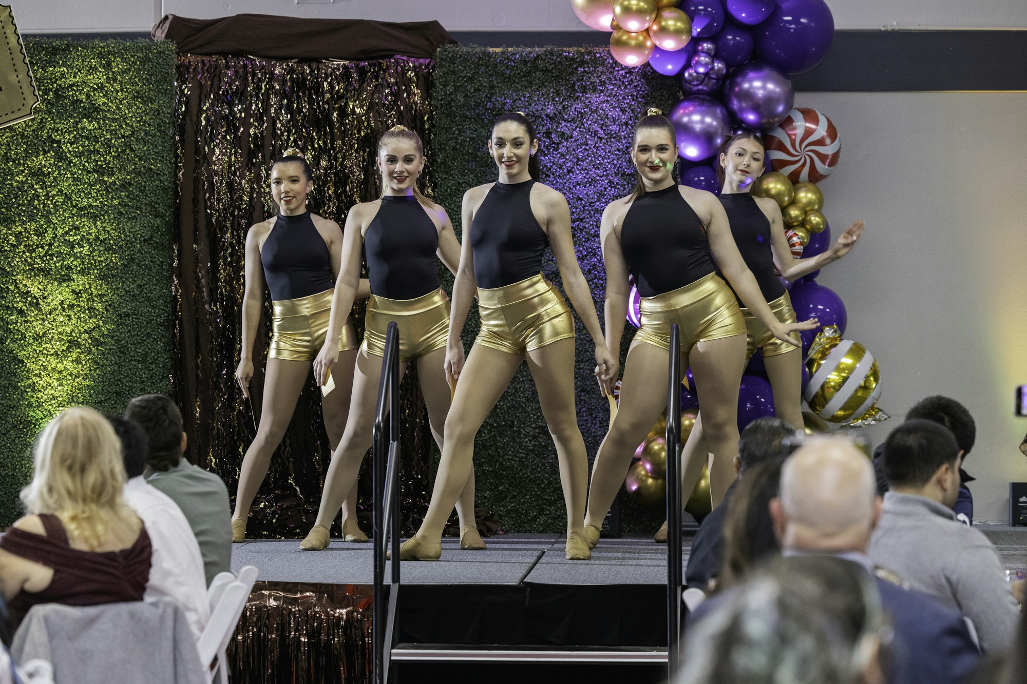 Five dancers in black tops and gold shorts perform on stage; with balloons and an audience in the background.
