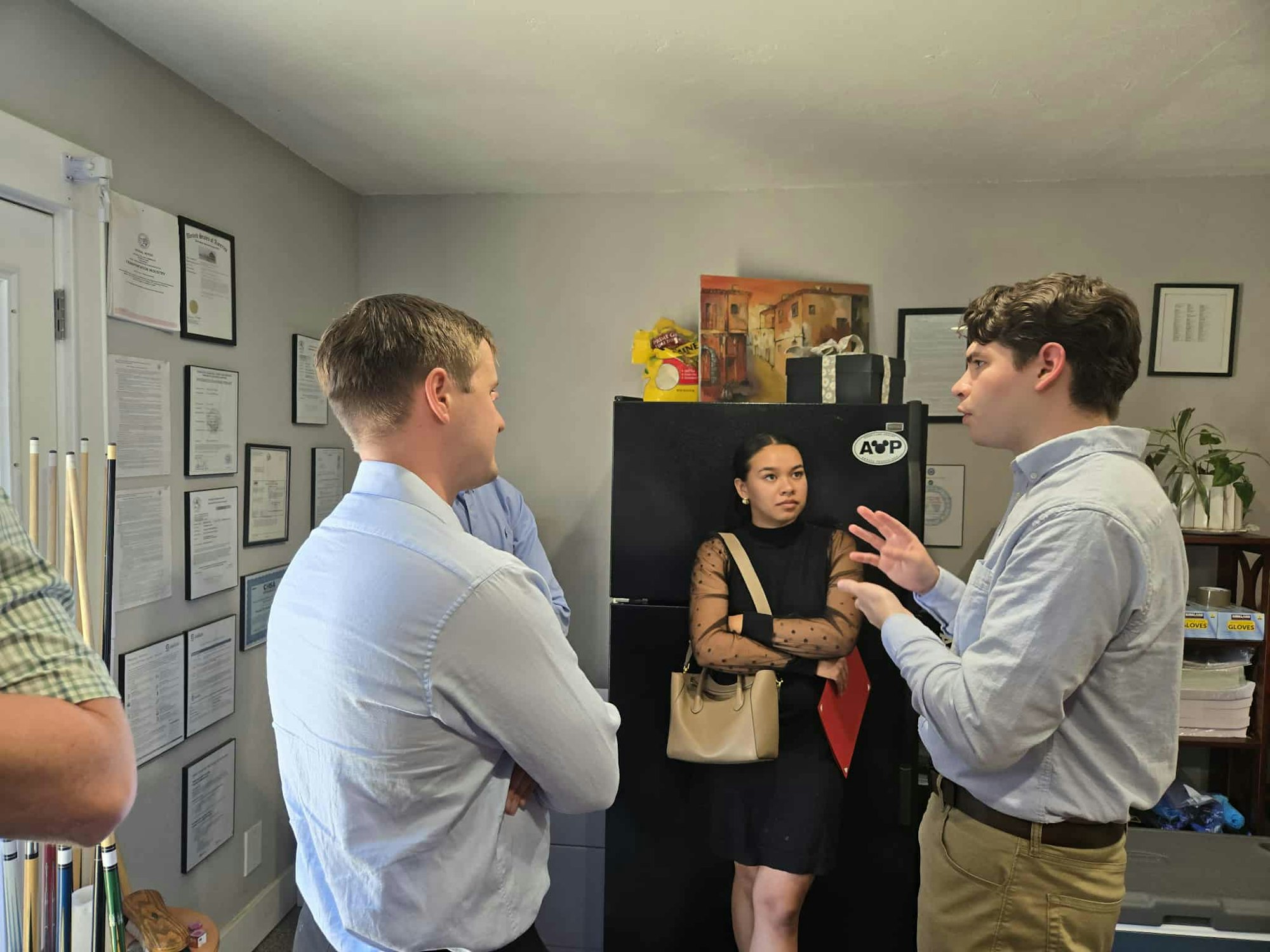 Four people in a room talking, with framed certificates on the wall and a fridge in the background.