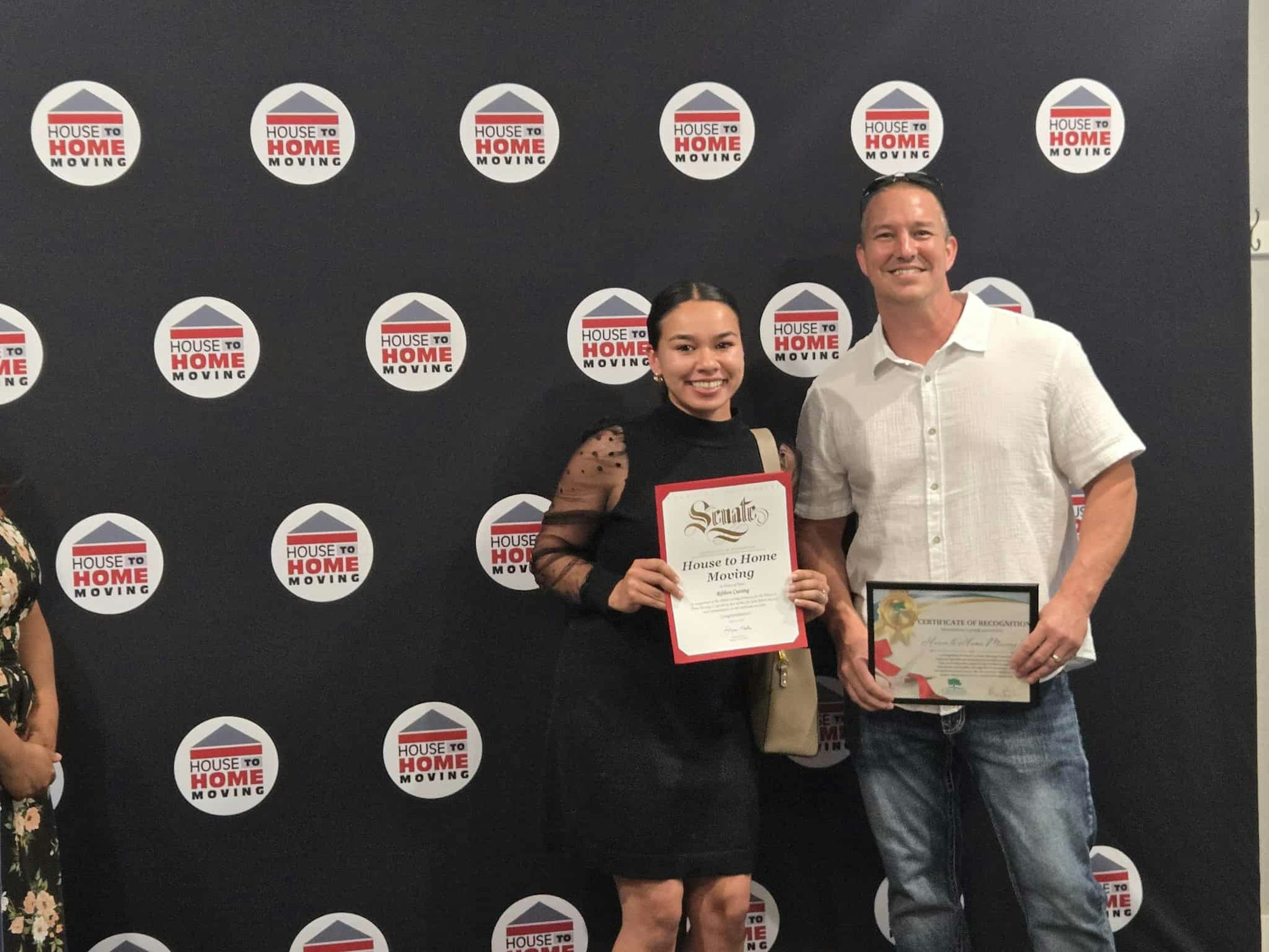 Two people holding certificates, standing in front of a backdrop with "House to Home Moving" logos.