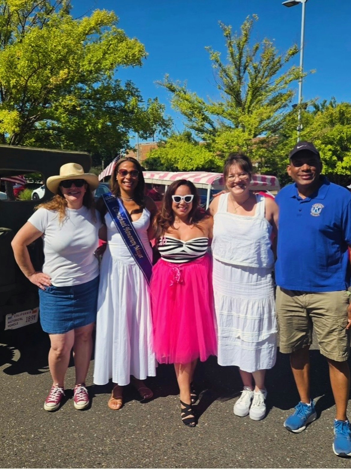 A group of five people posing outdoors, wearing colorful outfits, with trees and a clear blue sky in the background.
