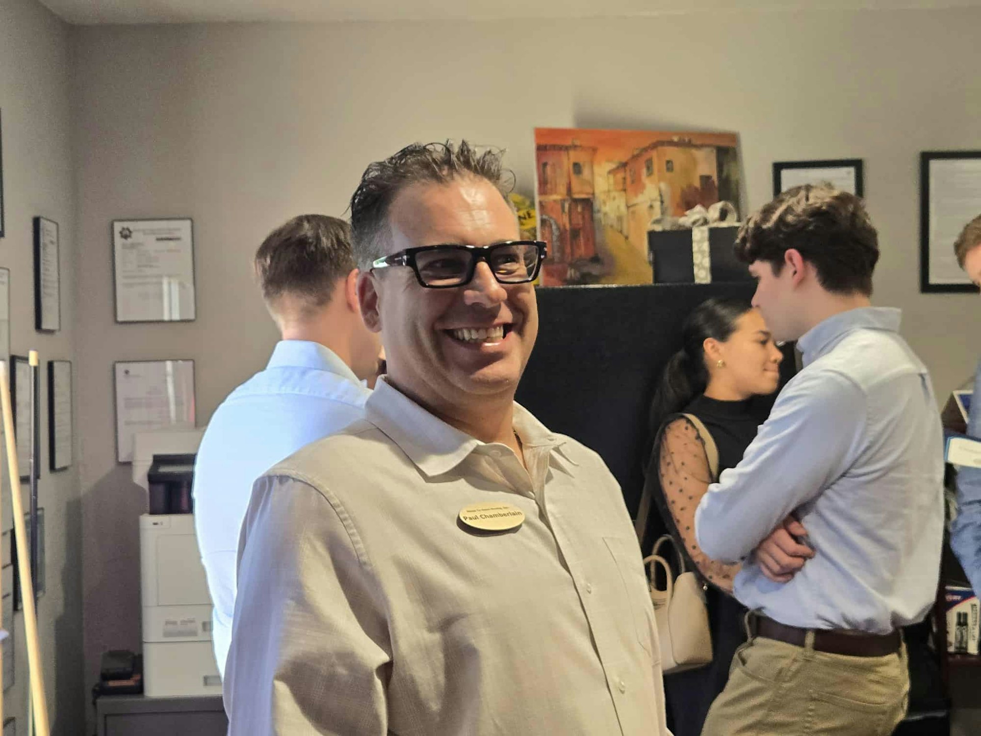 A group of people in a room, one person smiling in the foreground, others engaged in conversation in the background.