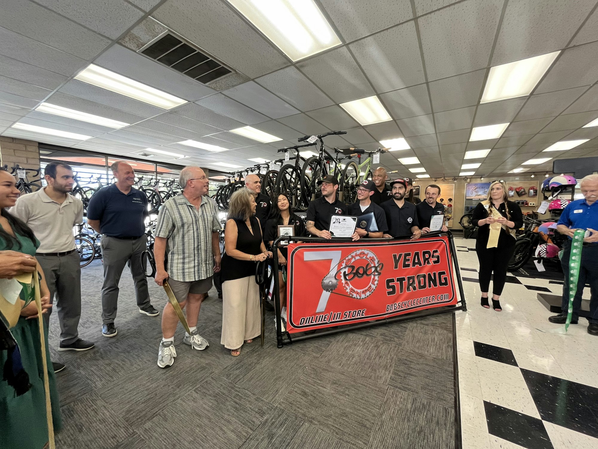 A group of people in a bicycle store celebrating "70 Years Strong" with a banner and certificates, indoors.