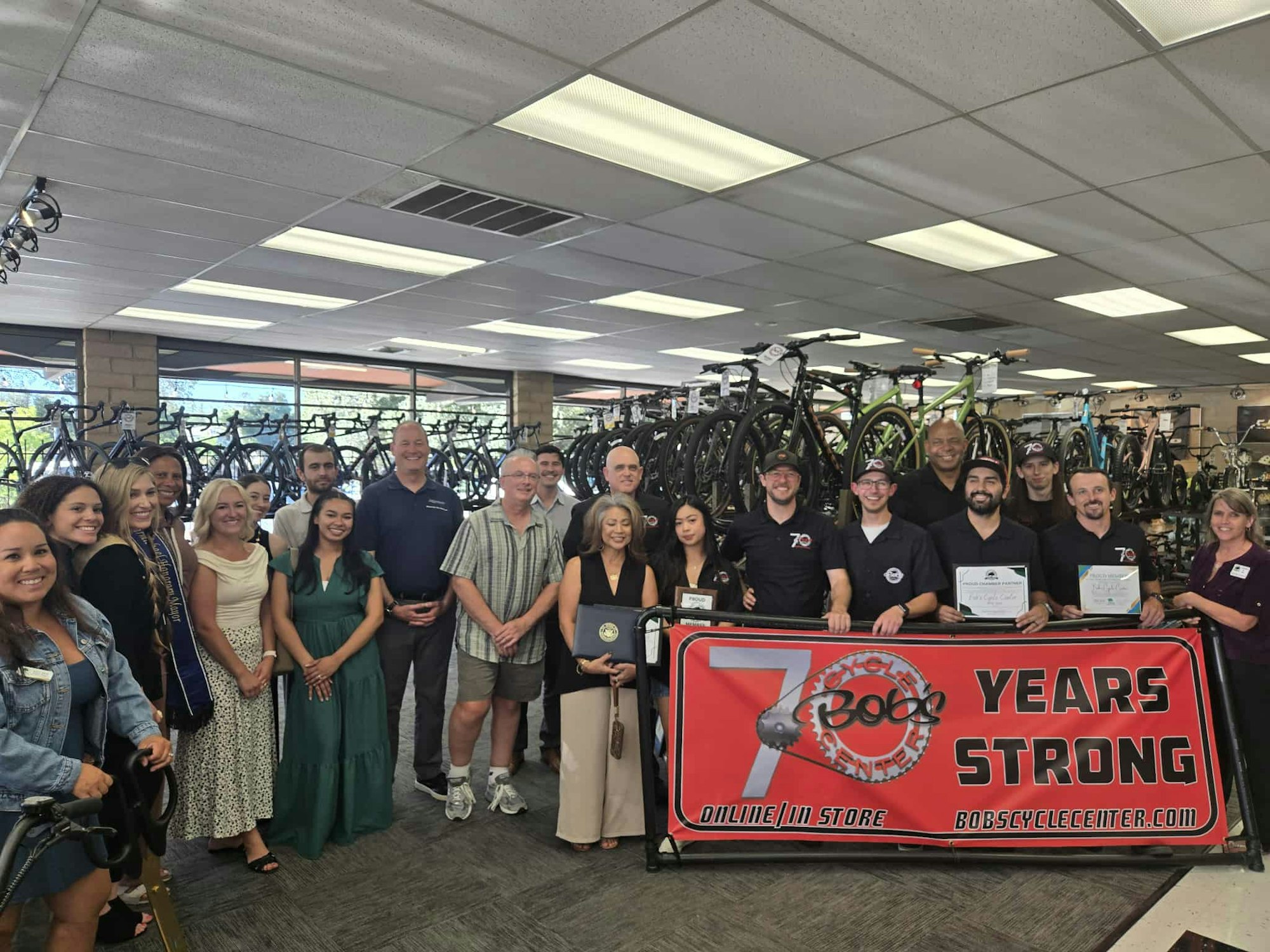 A group of people in a bicycle store celebrating "70 Years Strong," with a banner and certificates.