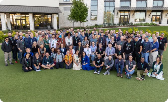 A large group of people posing together on a lawn outside a building, some seated and others standing, wearing conference badges.