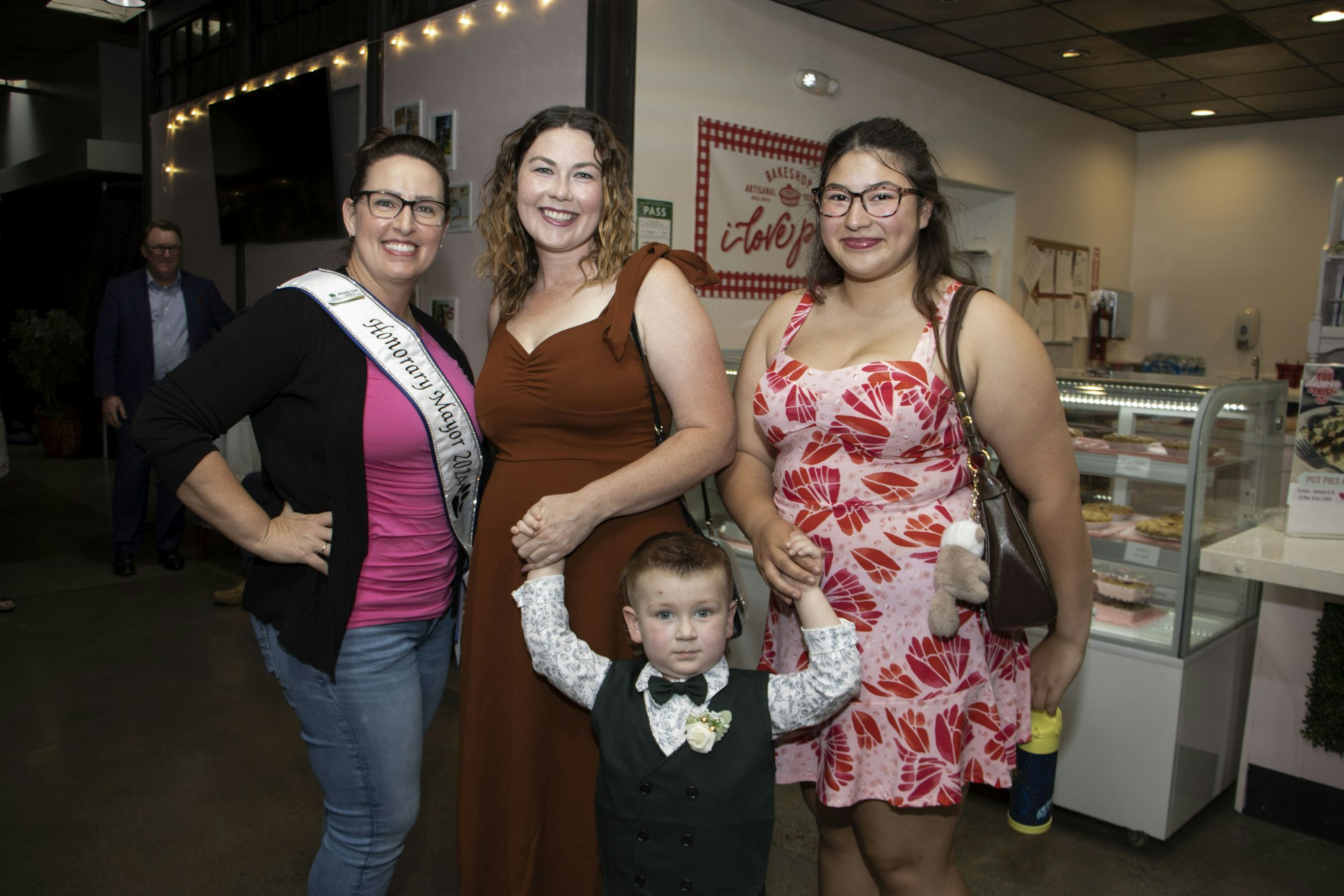 Three adults and a child posing, one wearing a sash with "Honorary Mayor 2018", inside a shop with pastries on display.