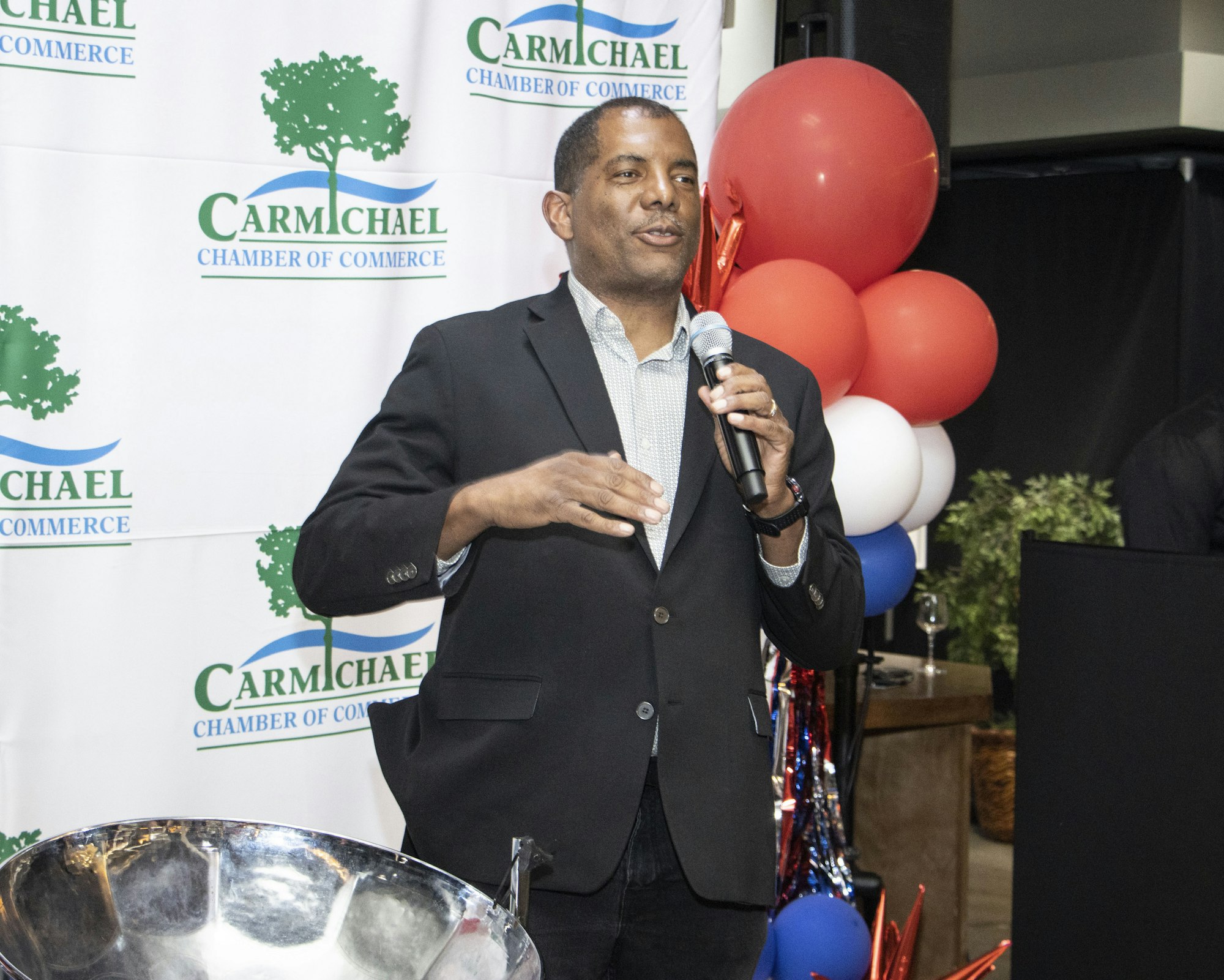 A man speaking at a Carmichael Chamber of Commerce event, with a microphone and decorations in the background.