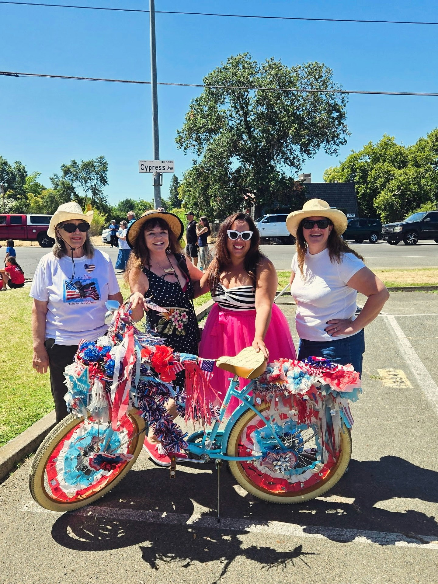Four women in festive clothing stand by a bicycle decorated with red, white, and blue streamers, likely celebrating a patriotic event.
