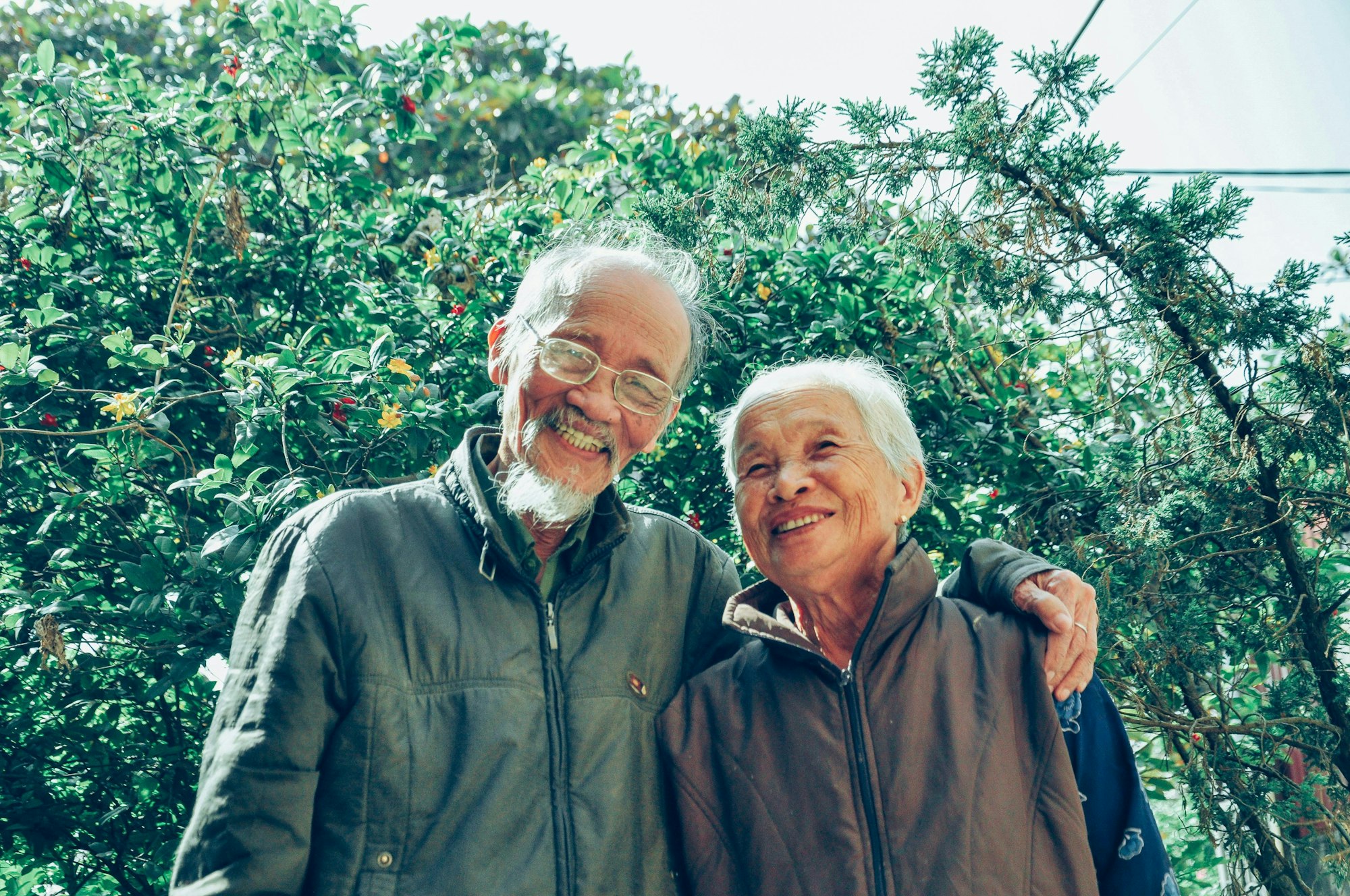 Two smiling elderly people standing together outdoors, surrounded by greenery.