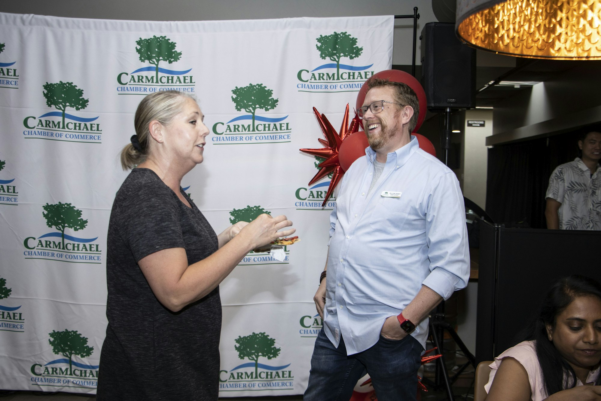Two people converse at a Carmichael Chamber of Commerce event, with a branded backdrop and a festive atmosphere.