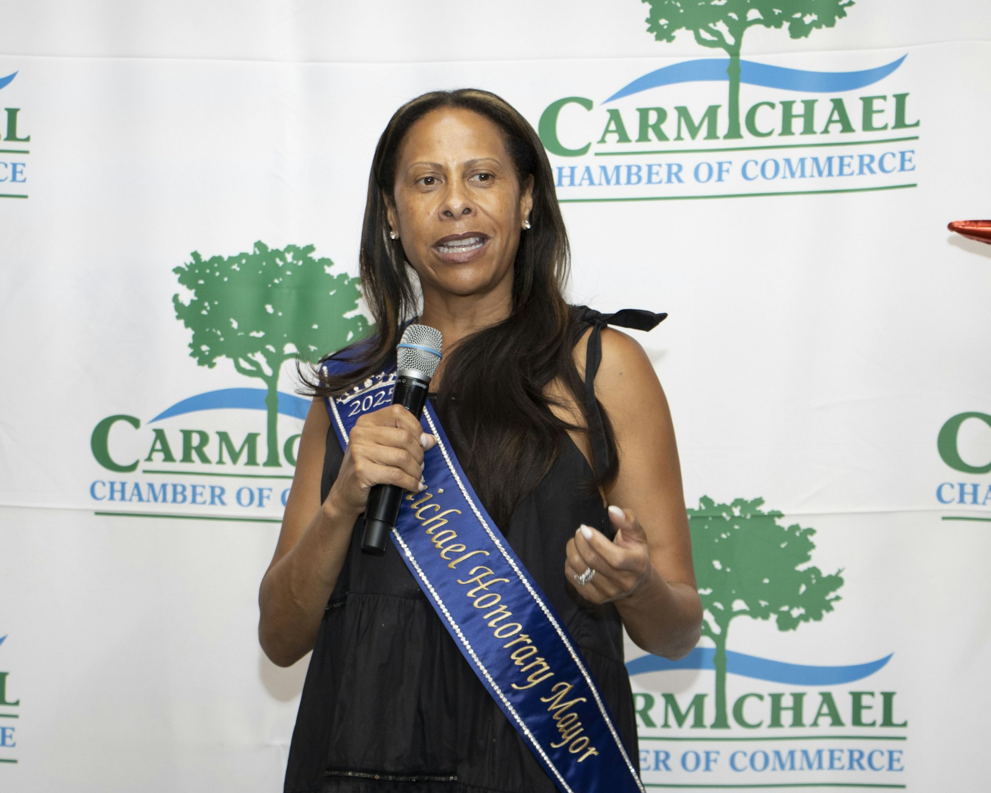 A person speaking with a microphone, wearing a "Carmichael Honorary Mayor" sash, stands in front of a "Carmichael Chamber of Commerce" banner.