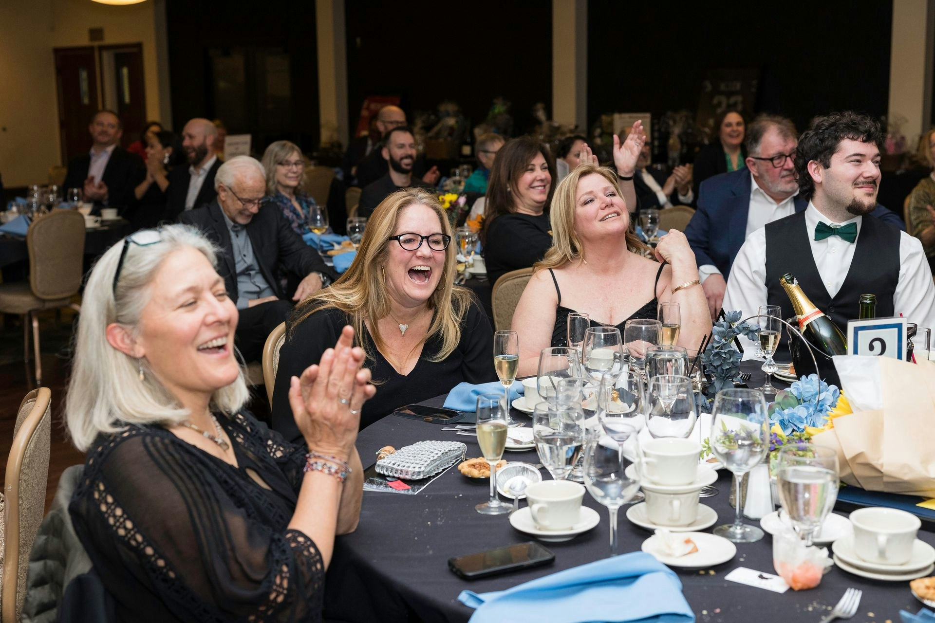 People at a banquet table are clapping and laughing, with dinnerware and glasses in the foreground.