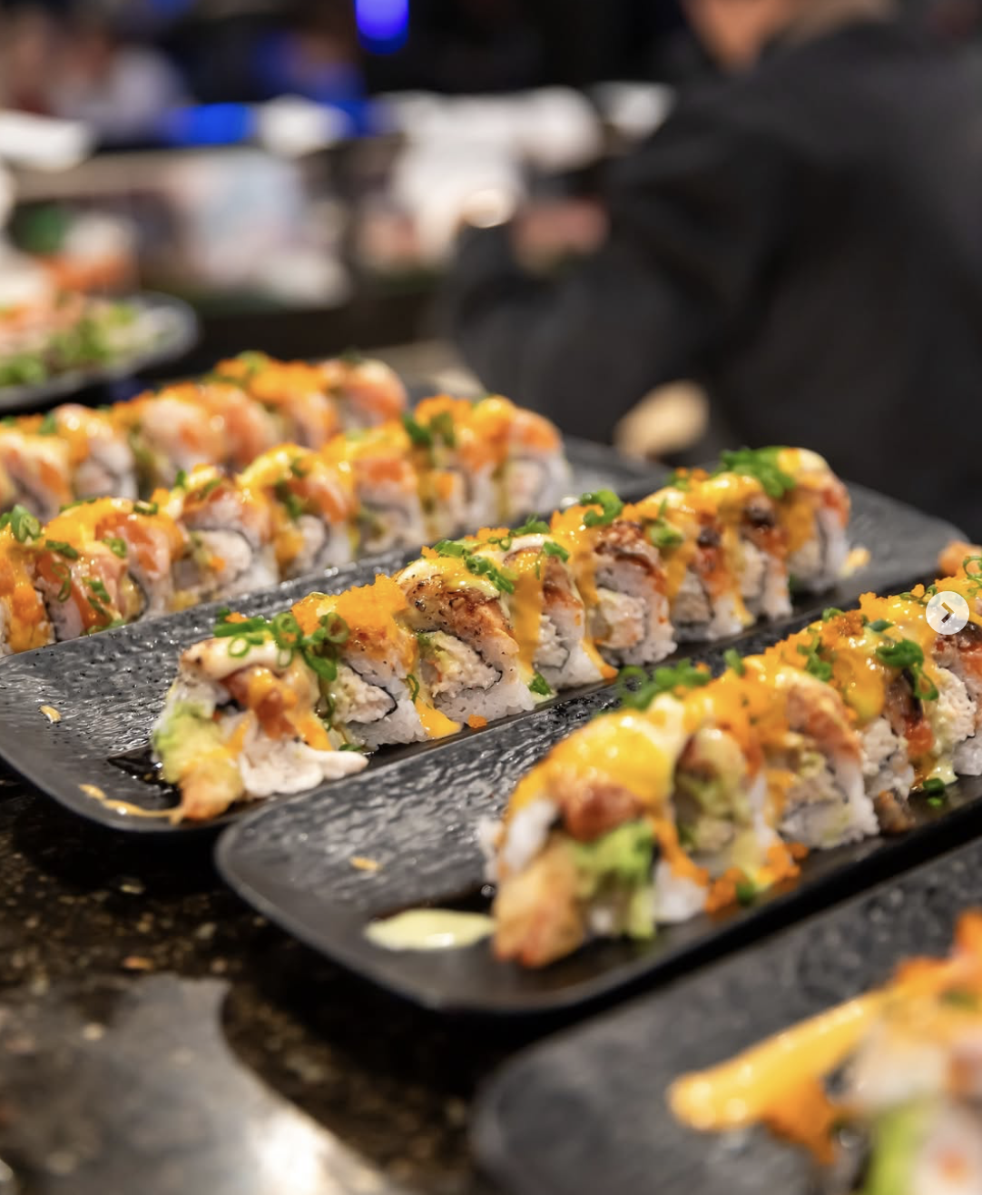 Sushi rolls on black plates, topped with sauces and garnishes, arranged neatly in rows.