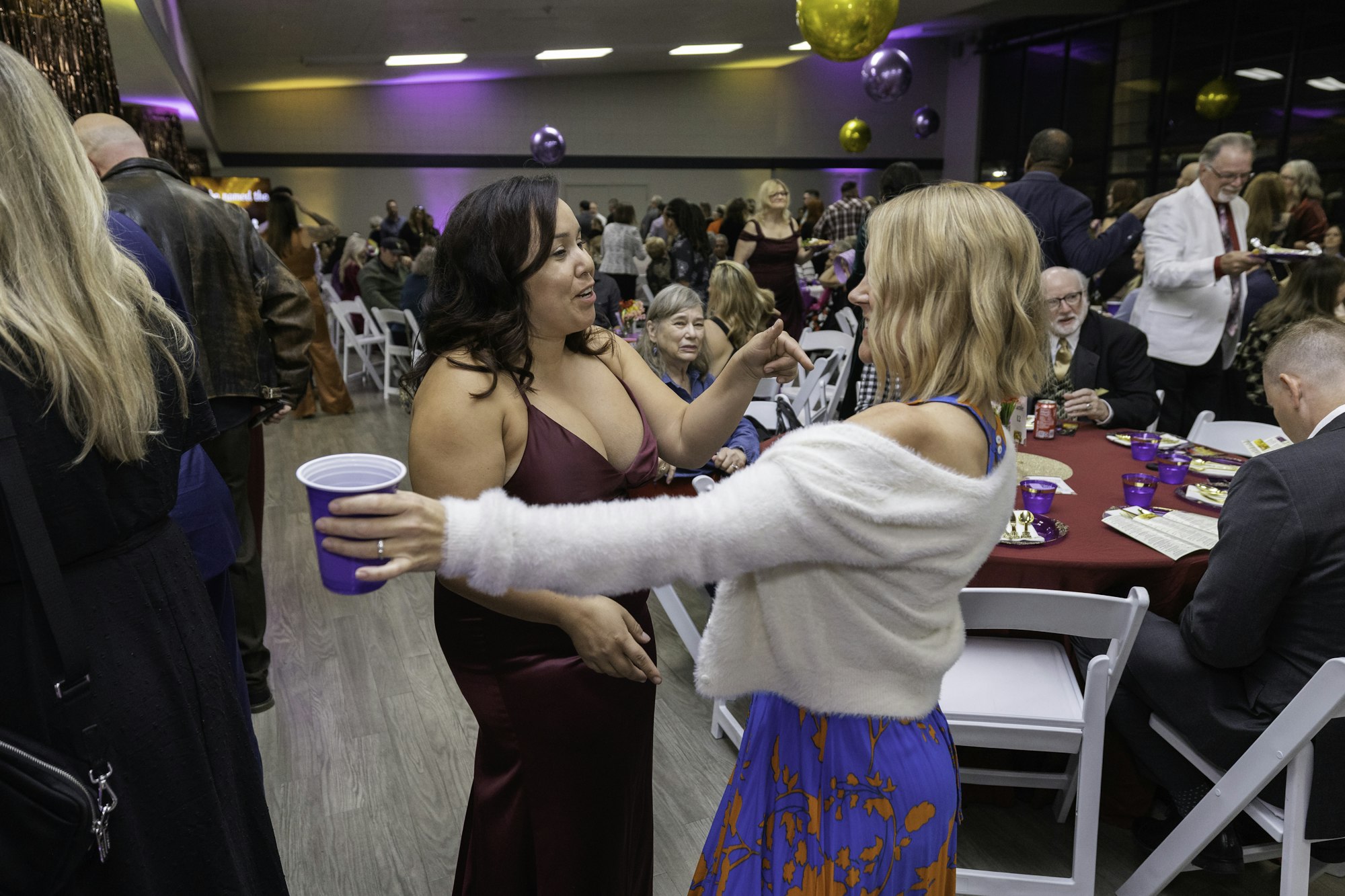 People socializing and dancing at a crowded indoor event with purple lighting and party decorations.