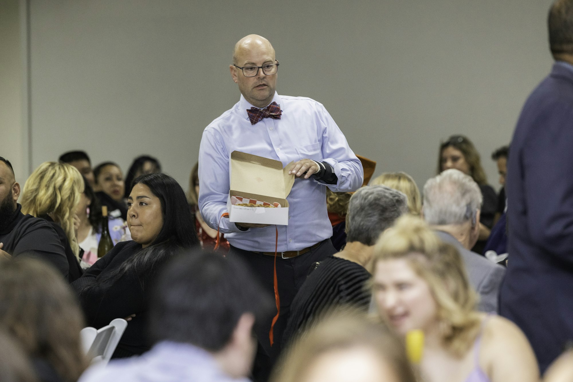 A man in a bowtie stands in a crowded room, holding an open box.
