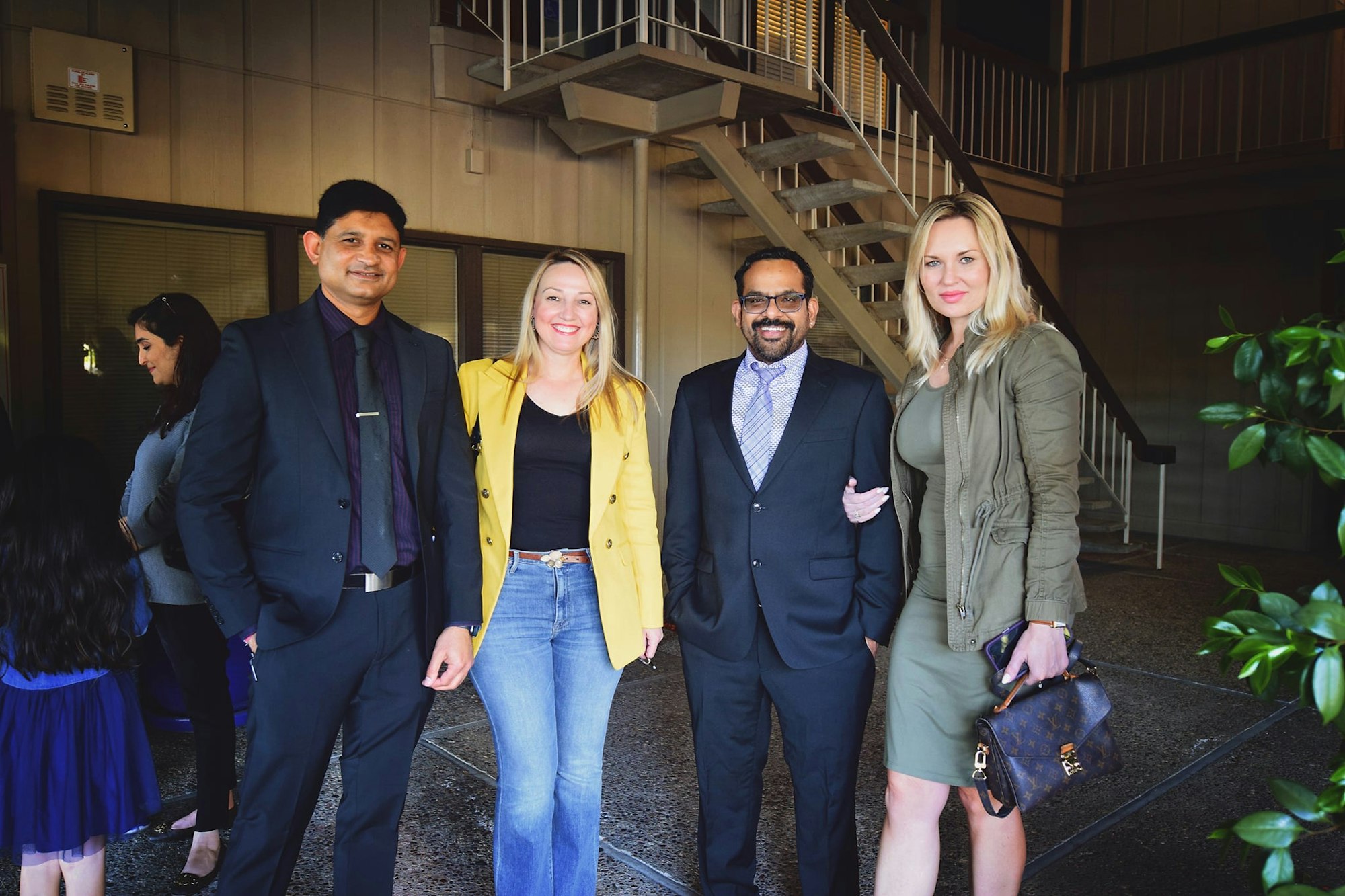 Four people posed in a semi-formal setting near stairs, with others in the background.