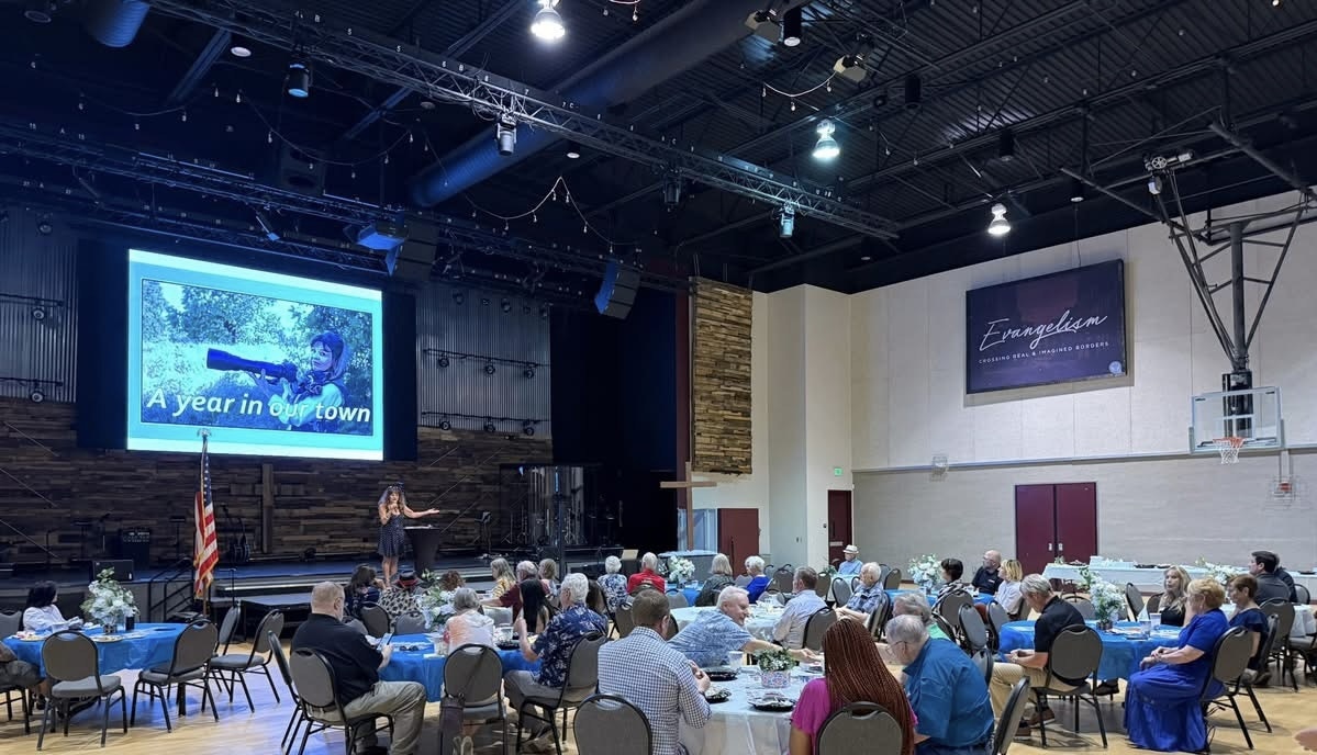 Audience at an indoor event with a speaker on stage, a "A year in our town" presentation slide, and tables with people.