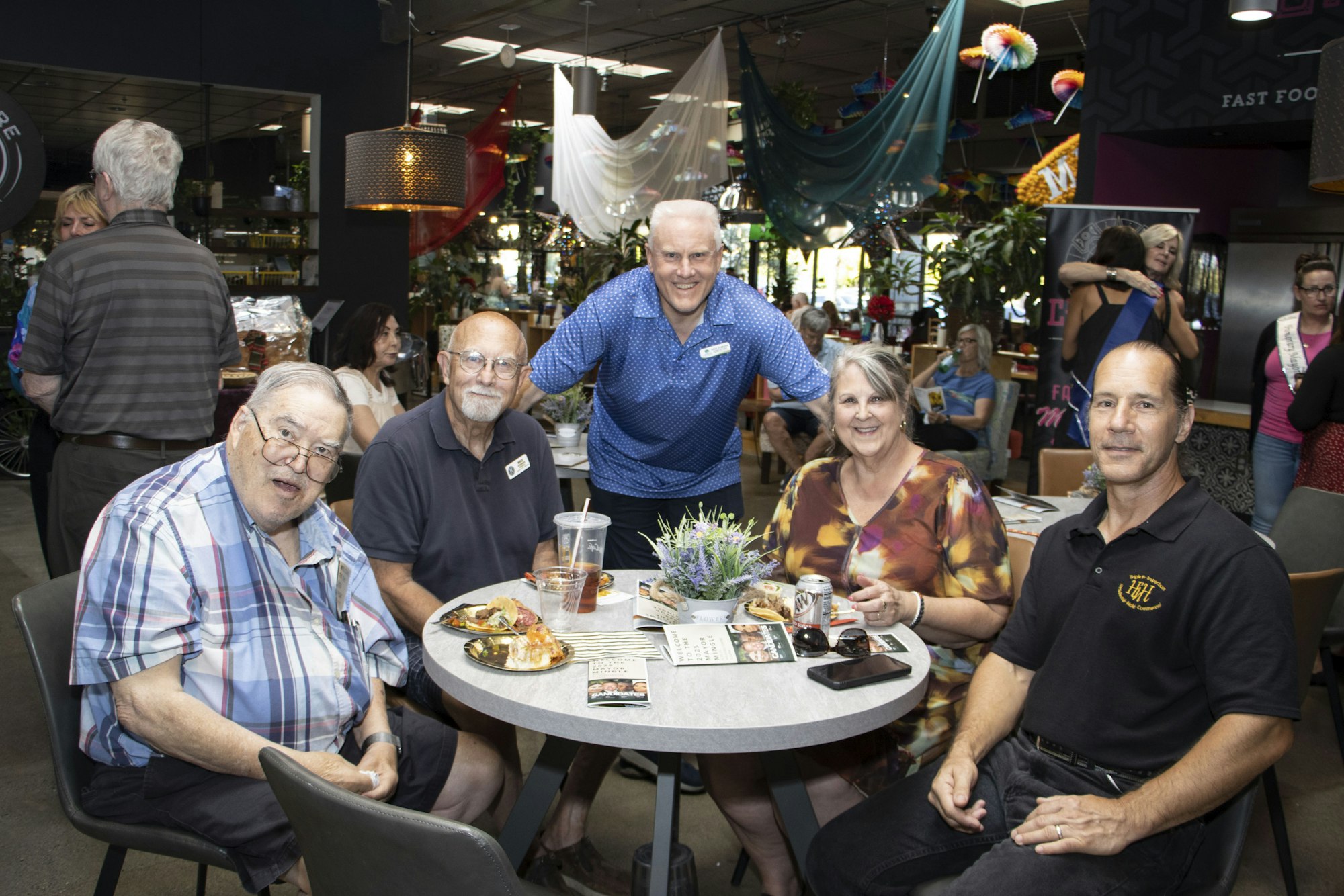 A group of people sitting around a table with food and drinks, inside a lively setting with colorful decorations and others in the background.