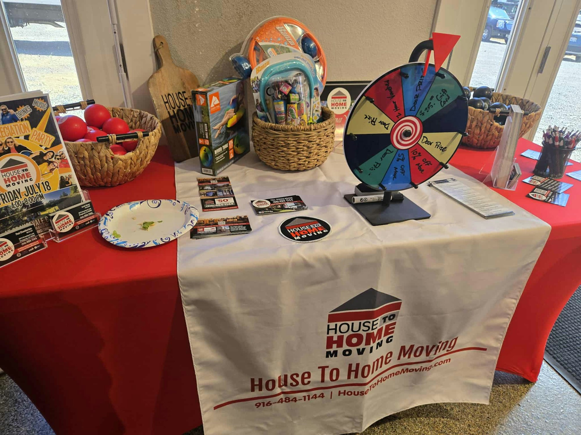 A promotional table with a prize wheel, flyers, baskets of items, and a House To Home Moving banner.
