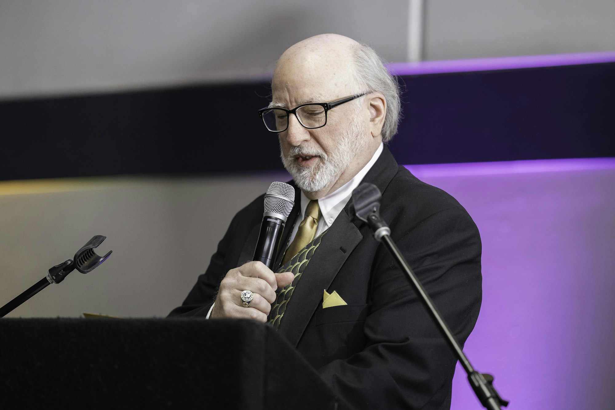 A man in a suit and glasses is speaking into a microphone at a lectern.